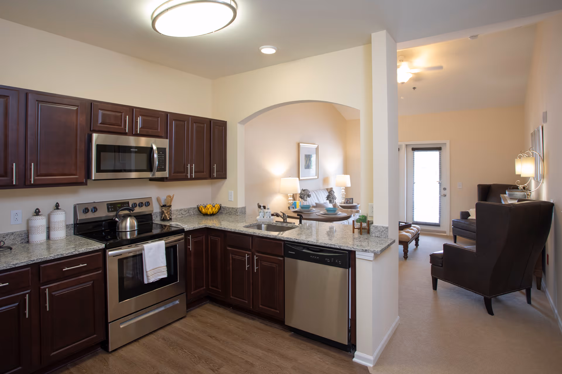Open-plan kitchen with dark wood cabinets, stainless steel appliances and granite countertops opening to a living area.