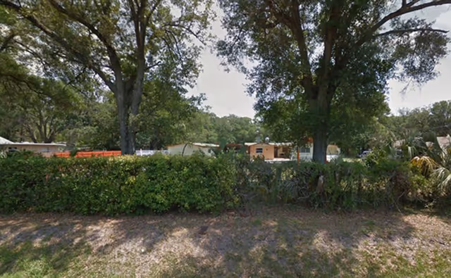 Outdoor view of a facility named Angels Garden Aclf, showing a green hedge and several large trees in the foreground with single-story buildings partially visible behind the trees under a partly cloudy sky.