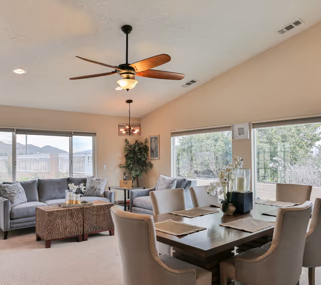 Open, airy living and dining area with a wooden dining table and upholstered chairs, gray sofas, large windows, and a ceiling fan.