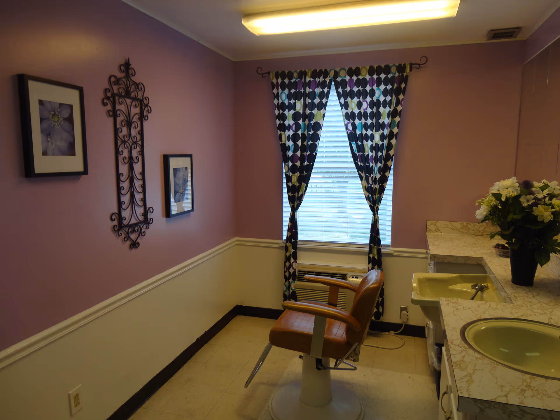 A small room with lavender walls and a white lower wall panel. There is a window with patterned curtains and blinds, a brown salon-style chair in front of a countertop with two sinks, and a vase of flowers on the counter. The wall has two framed pictures and a decorative wrought iron piece.