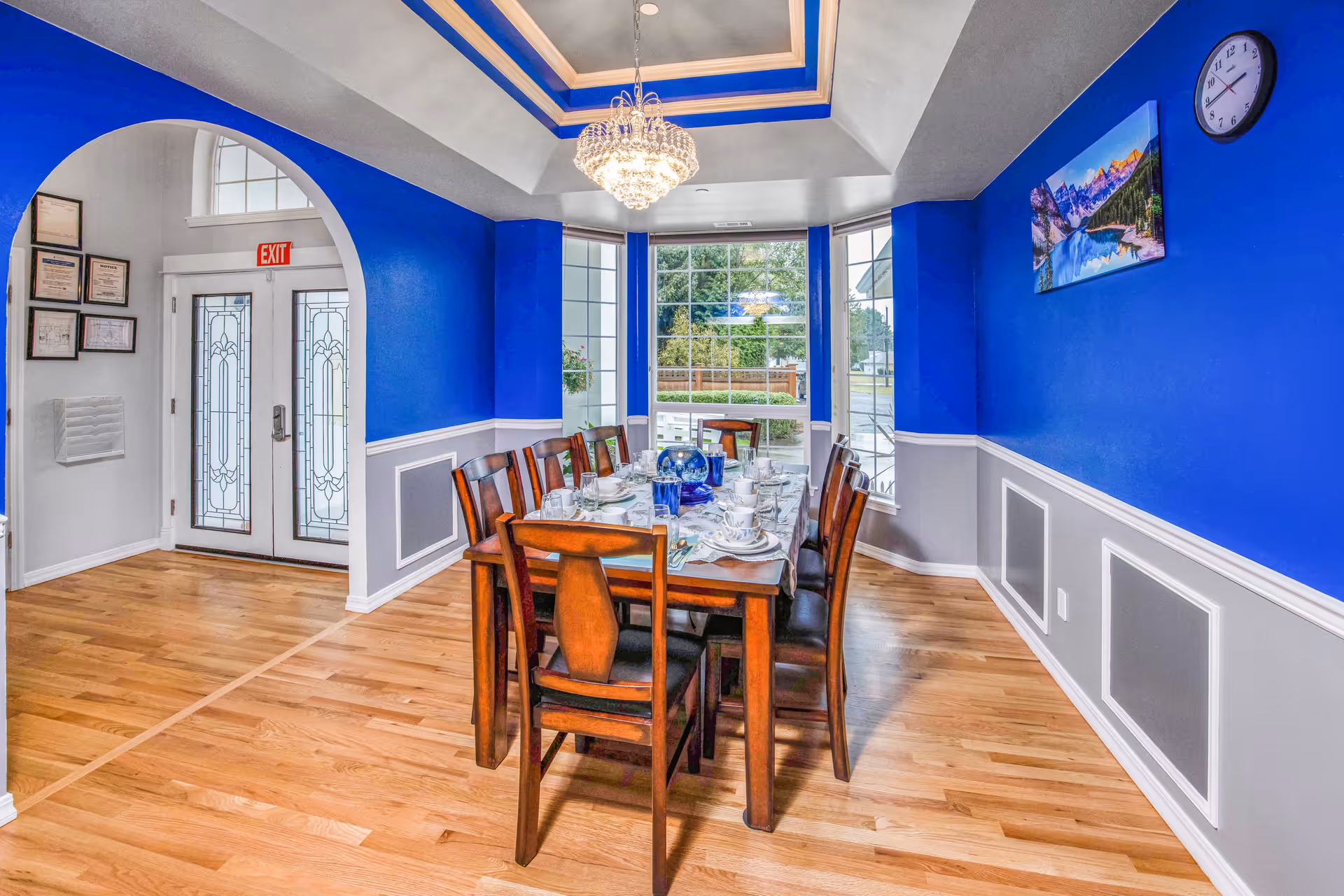 A dining room with bright blue and gray walls, a wooden dining table set for eight with dishes and glassware, a large window with a view of greenery outside, a chandelier hanging from a tray ceiling, a wall clock, and a landscape painting on the wall.