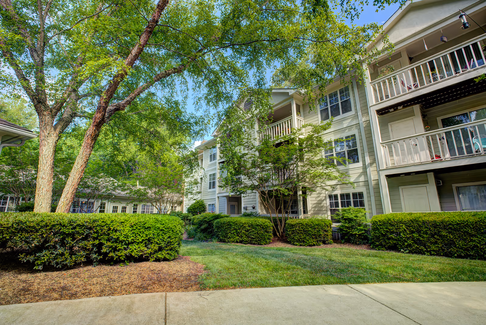 Exterior view of a multi-story senior living building with balconies, mature trees, hedges, and a landscaped courtyard.