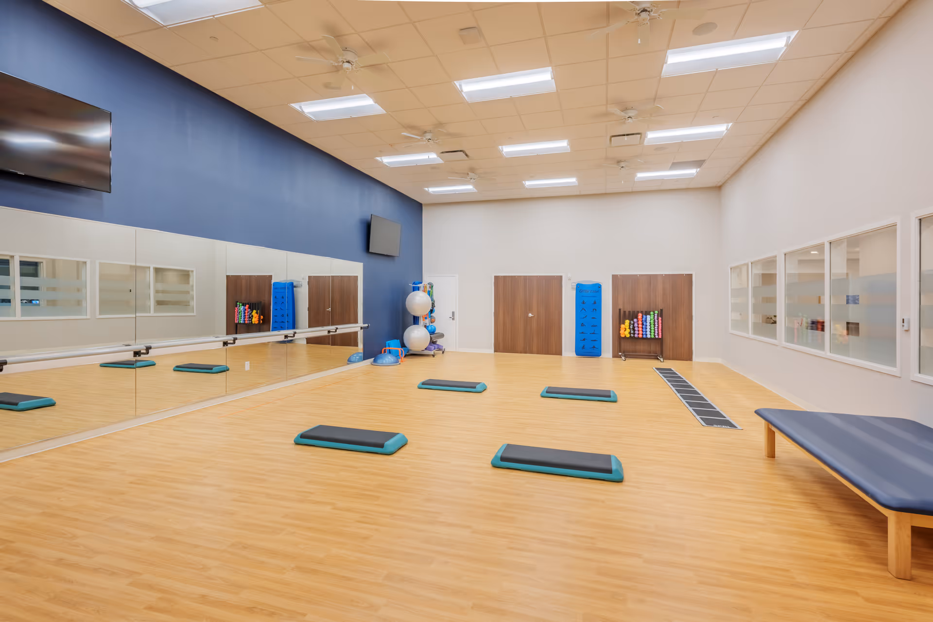A spacious exercise room with wooden flooring and a blue accent wall. The room contains several exercise steps arranged on the floor, a bench on the right side, and various fitness equipment such as exercise balls and weights stored against the far wall. Large mirrors cover one wall, and two flat-screen TVs are mounted on the walls. The ceiling has multiple fluorescent lights and ceiling fans.