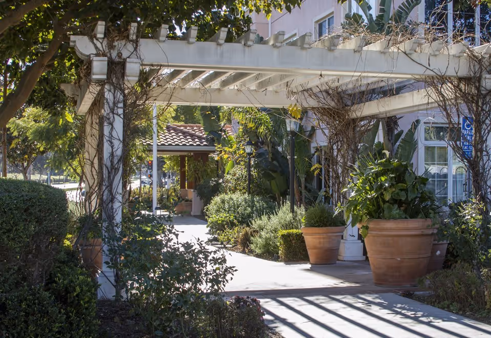 Outdoor walkway at Ivy Park at Playa Vista featuring a white pergola with climbing plants, large potted plants, and lush greenery along the path leading to a building entrance with a tiled roof.