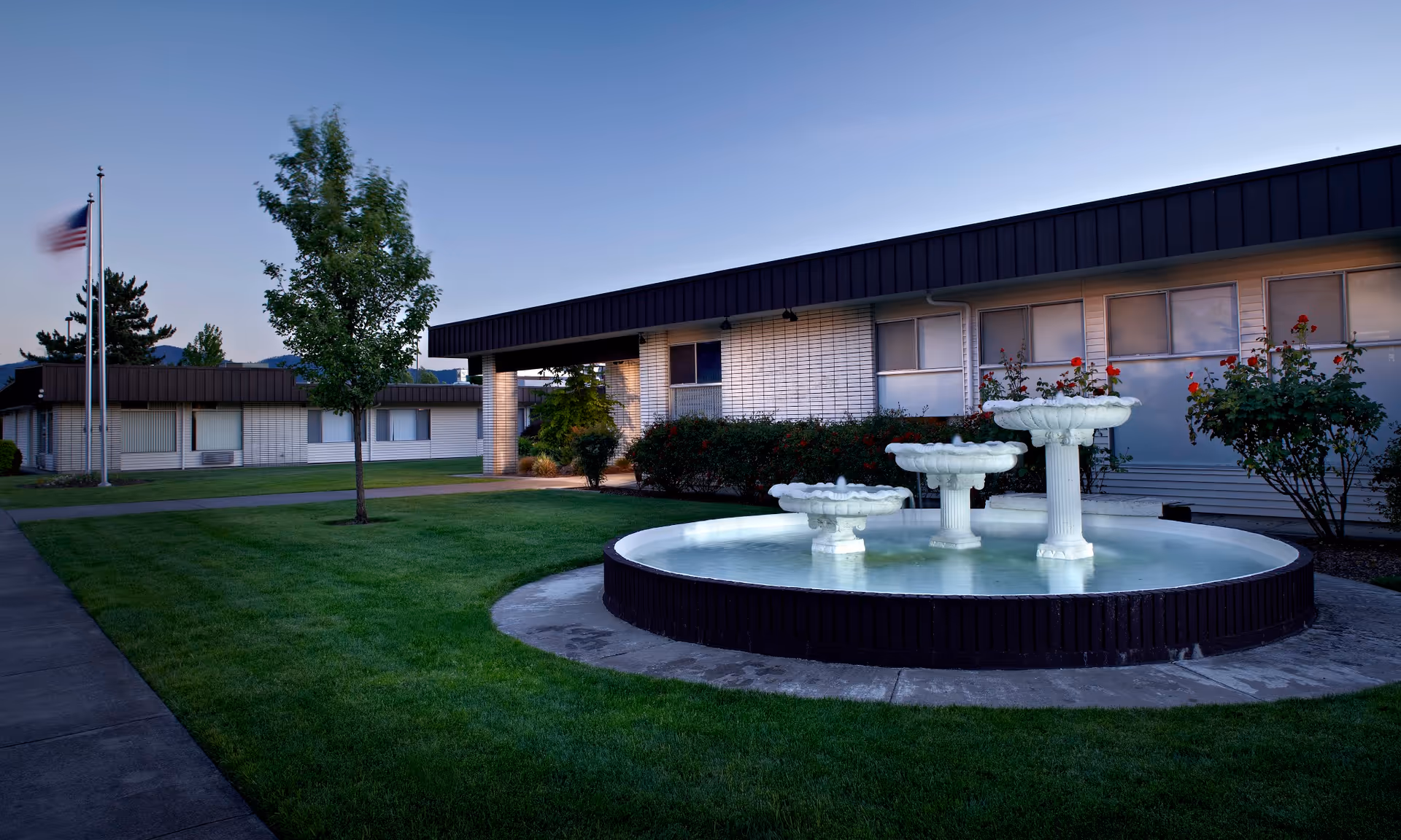 Outdoor view of a senior living facility with a circular water fountain in the foreground, green grass, a tree, and a building with white brick walls and a dark roof in the background during dusk.