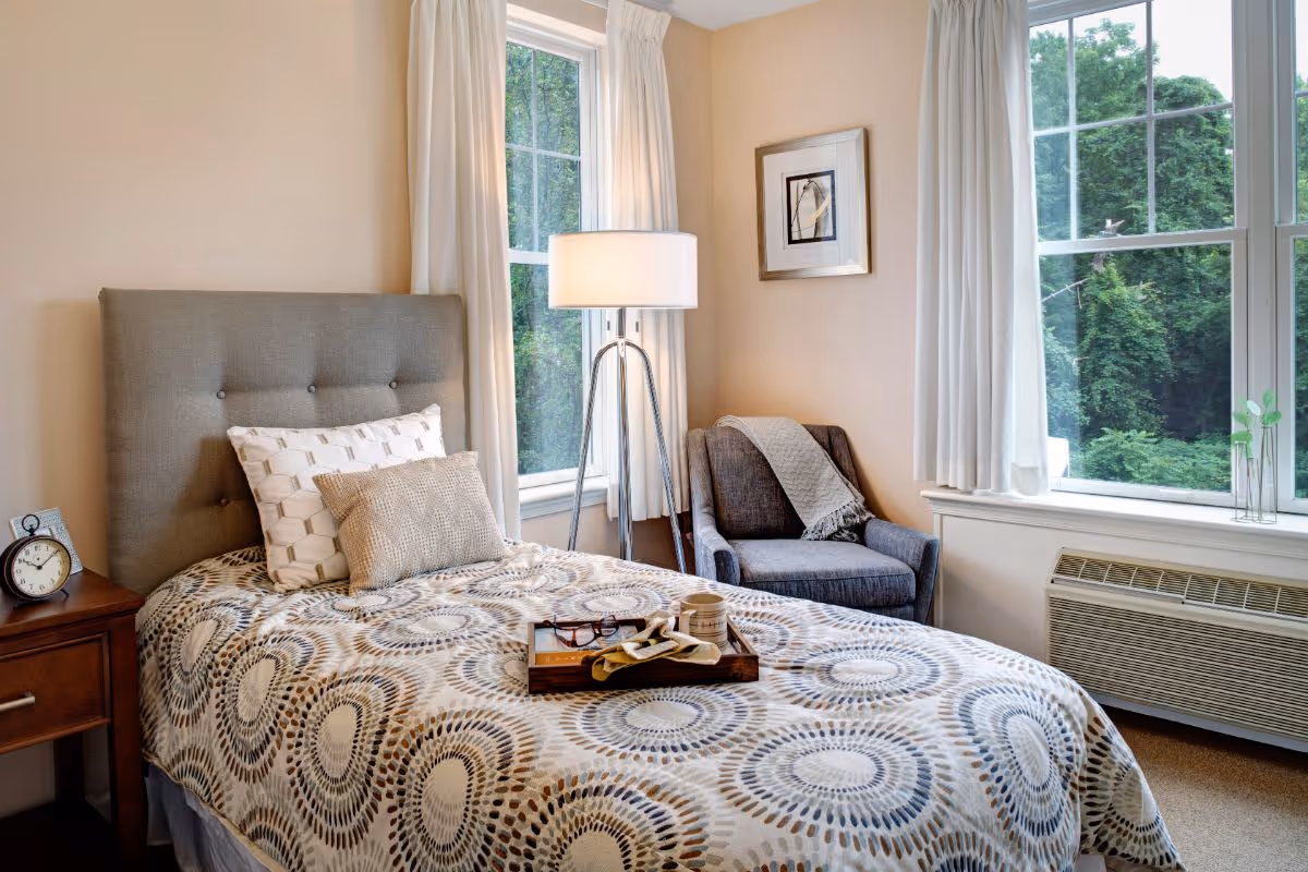A cozy bedroom with a single bed featuring a patterned bedspread and two pillows. Next to the bed is a wooden nightstand with an alarm clock and a framed photo. A floor lamp stands between two large windows with white curtains, and a gray armchair with a throw blanket is positioned in the corner. Outside the windows, green trees are visible.