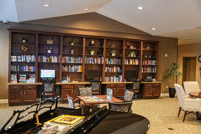 Community lounge with wooden bookshelves and computer stations, tables and chairs, and a piano in the foreground.