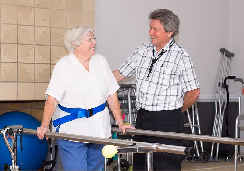 An elderly woman wearing a white shirt and blue pants is using parallel bars for physical therapy, supported by a blue gait belt. A man in a plaid shirt stands beside her, smiling and offering encouragement. In the background, there are crutches, walkers, and a large blue exercise ball.