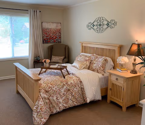A cozy bedroom with a wooden bed frame and a patterned bedspread. There is a wooden nightstand with a lamp, a book, and a flower vase. A cushioned armchair is placed near a window with curtains, and a decorative wall piece hangs above the bed.