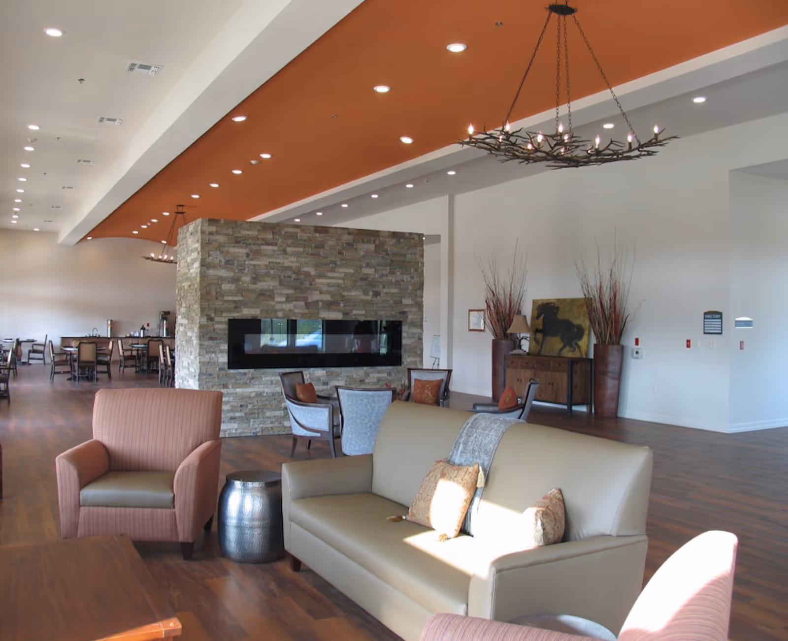 A spacious and well-lit living room area with a tan leather sofa, two upholstered armchairs, a small metallic side table, and a stone fireplace in the center. The ceiling features recessed lighting and an orange accent strip with a rustic chandelier. In the background, there are dining tables and chairs, a wooden sideboard with decorative items, and a painting of a horse on the wall.