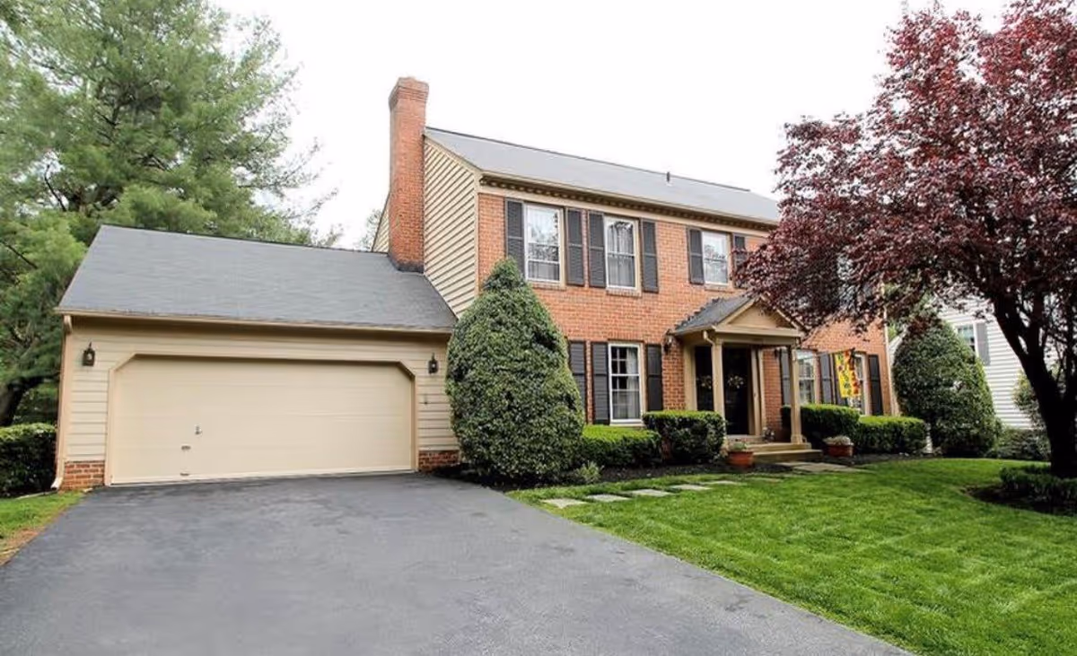 Front exterior view of a two-story brick and siding house with a double garage, a driveway, manicured lawn, shrubs, and a tree with reddish leaves.