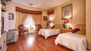 A warmly decorated bedroom with two single beds covered in beige quilts, each with a pillow. Between the beds is a wooden nightstand with a lamp and a small plant. The room has wooden flooring, two red cushioned chairs near a window with beige curtains and a red valance, and framed artwork on the walls. Soft lighting creates a cozy atmosphere.