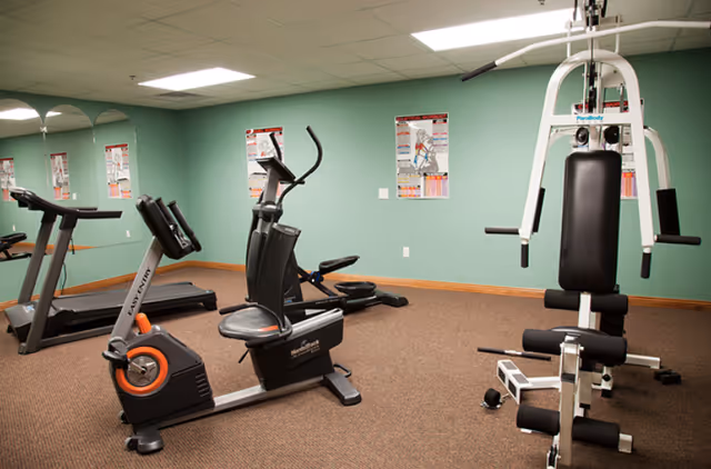 Exercise room with a stationary bike, treadmill, rowing machine and multi-station weight machine against green walls and mirrors.