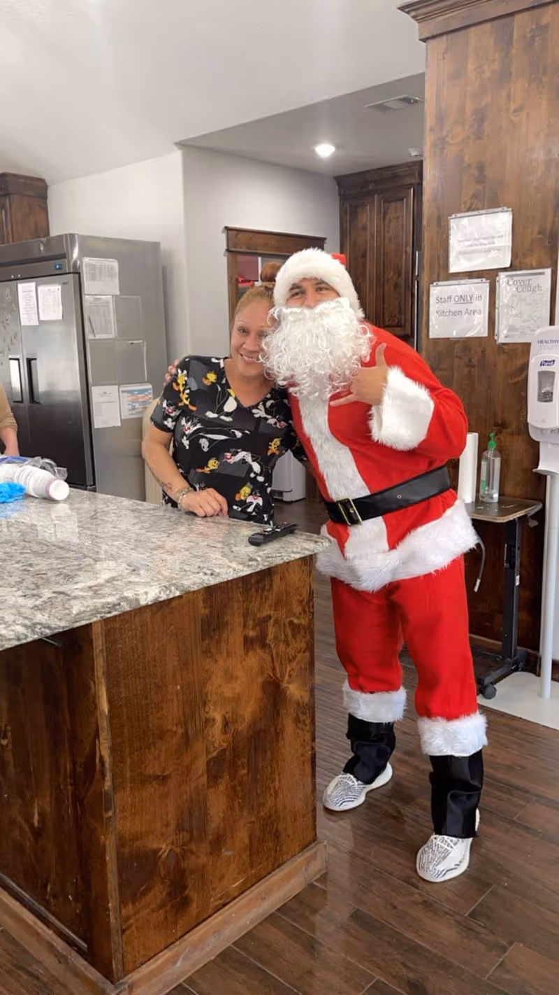 A woman standing behind a kitchen counter smiling and posing with a person dressed in a Santa Claus costume inside a kitchen area with wooden cabinets and a stainless steel refrigerator.