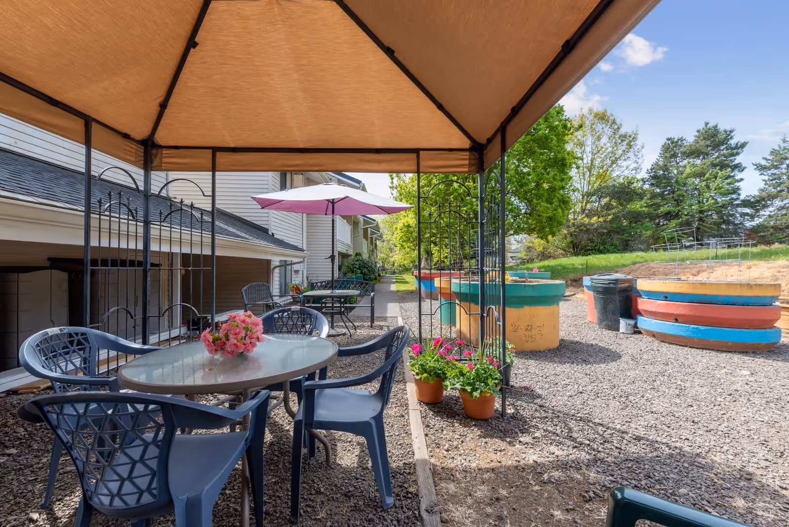 Outdoor seating area with a canopy overhead, plastic chairs around a glass table with a flower centerpiece, additional tables and chairs with umbrellas in the background, colorful raised garden beds, and green trees under a clear blue sky.