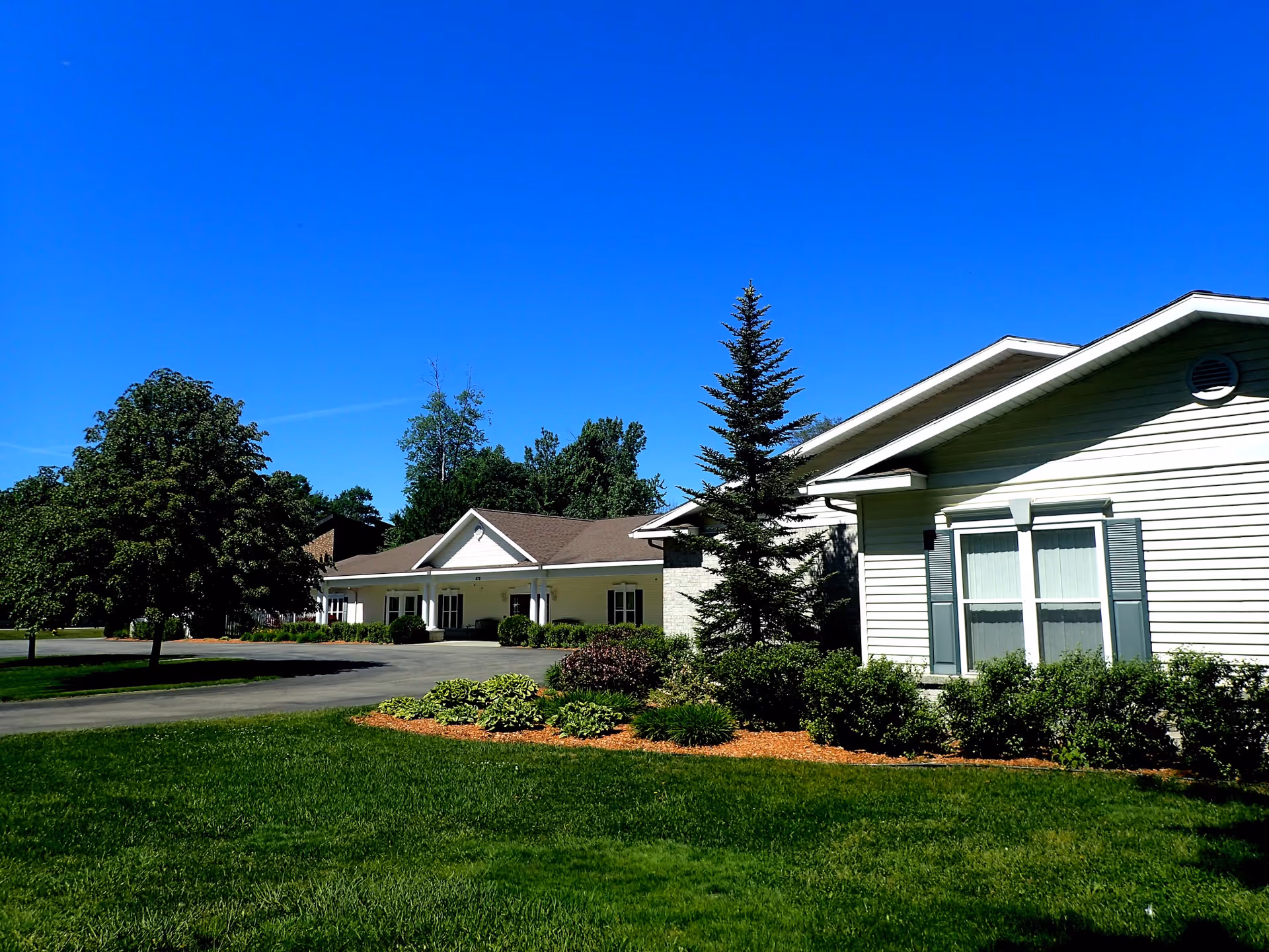 Front exterior of a single-story assisted living building with landscaped lawn, shrubs, and a clear blue sky.