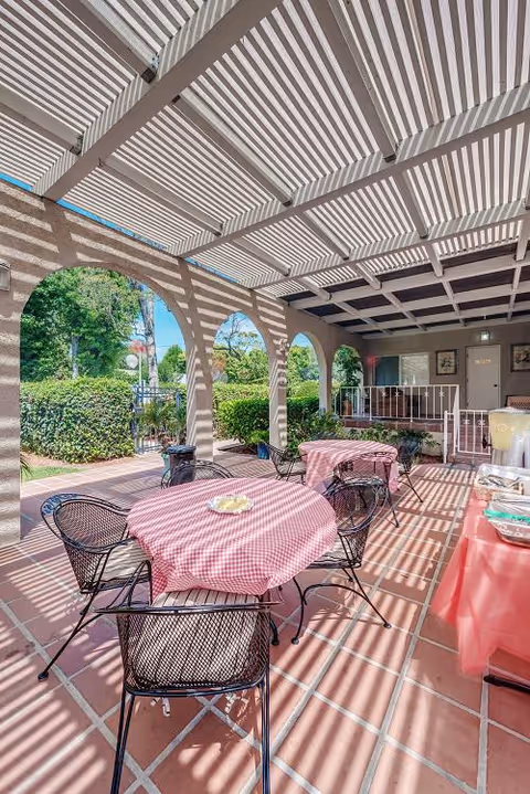 Outdoor covered patio area with arched openings and striped shadows from the overhead pergola. Several round tables with red and white checkered tablecloths and black metal chairs are arranged on a tiled floor. Green bushes and trees are visible outside the patio.