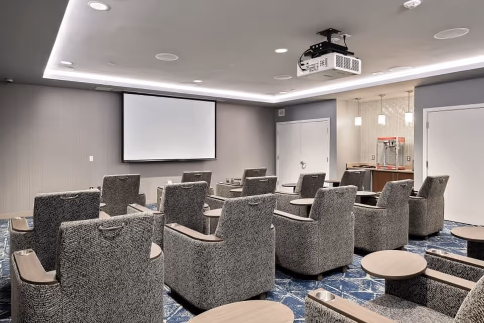 A modern media room with multiple gray upholstered armchairs arranged in rows facing a large white projection screen on the wall. The room has a ceiling-mounted projector, recessed lighting, and a popcorn machine in the corner near a small counter with pendant lights above. The floor is covered with a blue patterned carpet.