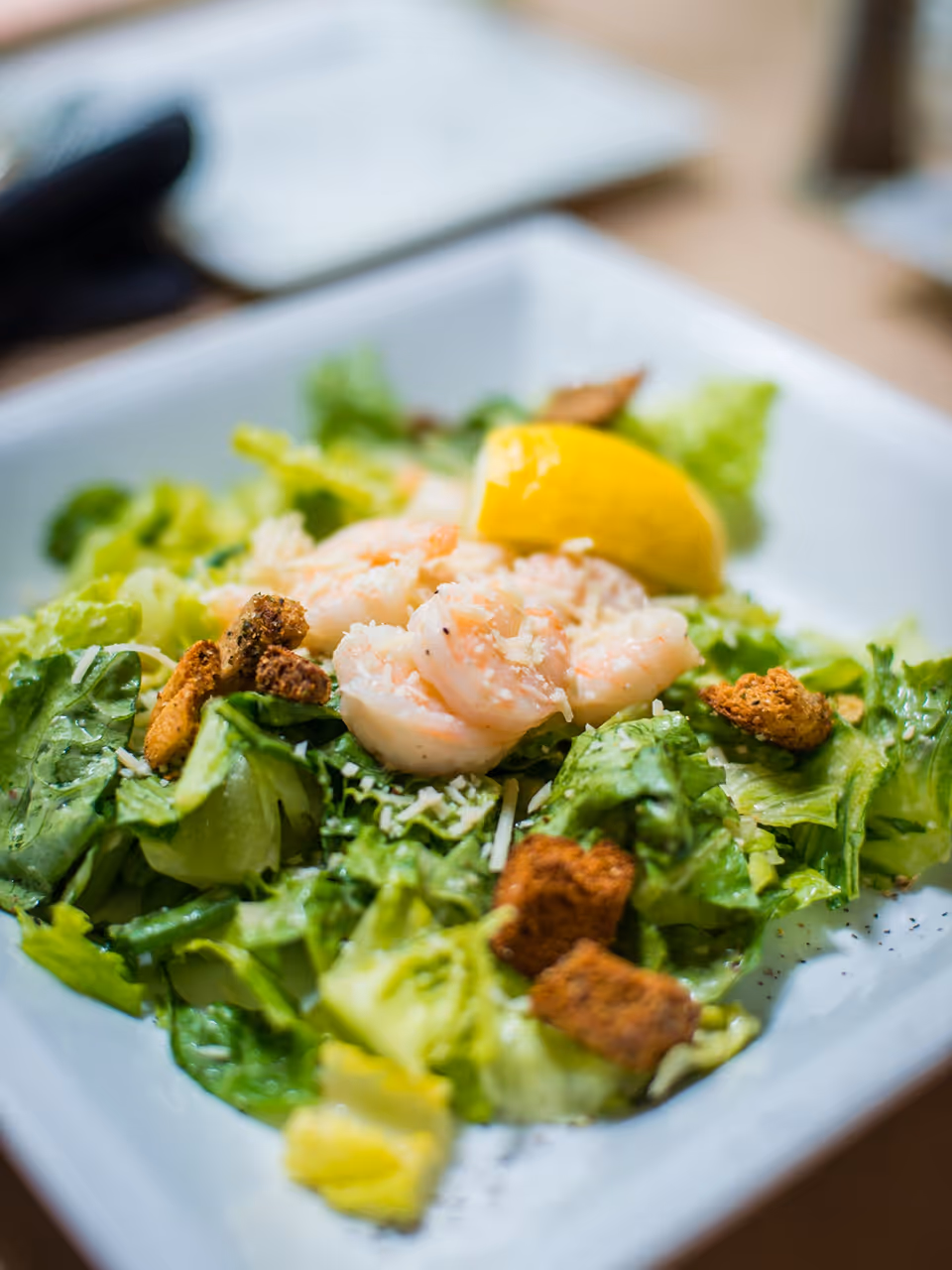 A close-up of a fresh salad with leafy greens, croutons, grated cheese, several cooked shrimp, and a lemon wedge on a white square plate.
