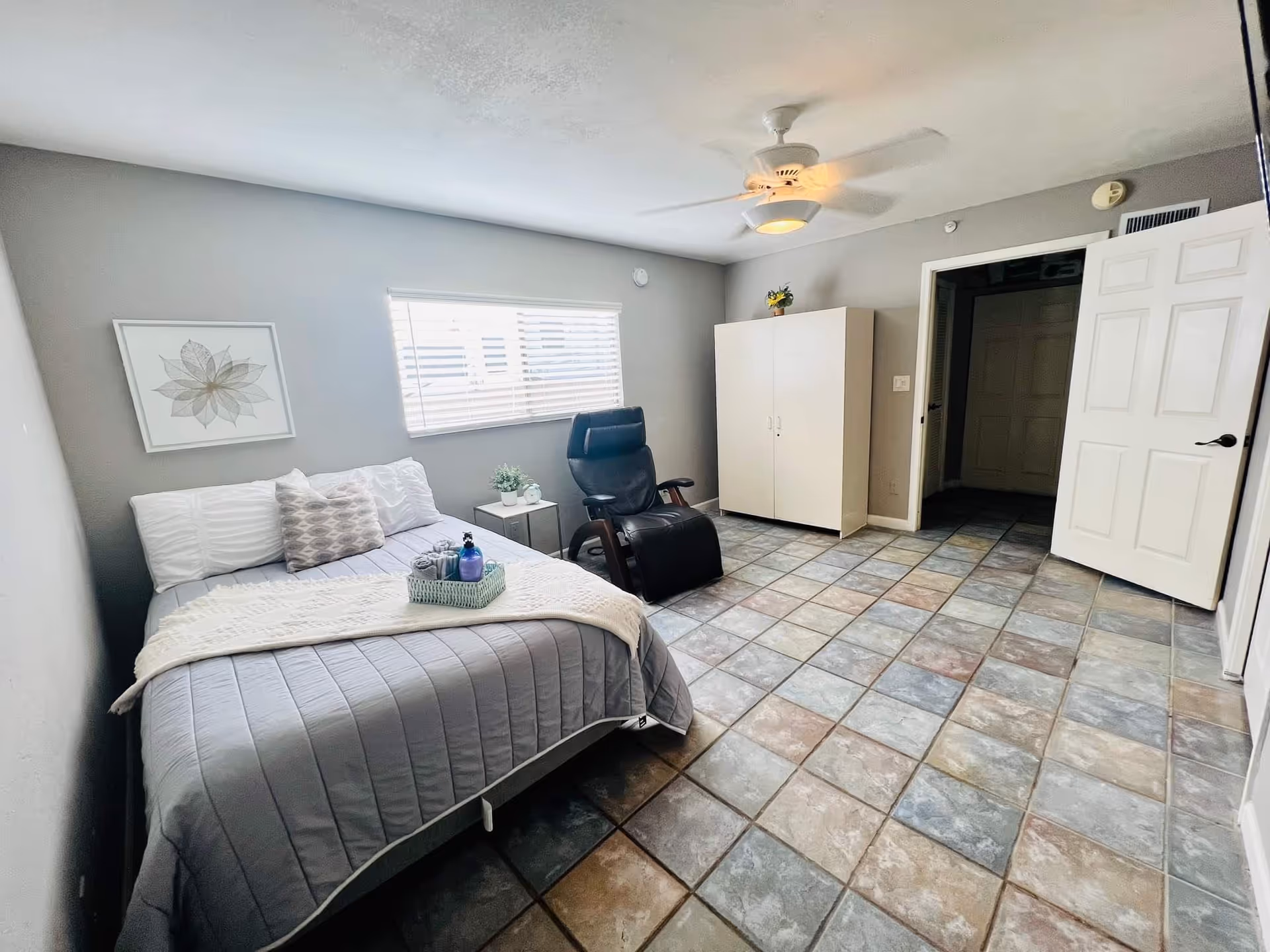 A bedroom with a bed covered in gray bedding and white pillows, a small side table with a plant and clock, a black recliner chair, a white wardrobe, and a ceiling fan. The room has tiled flooring and a window with blinds letting in natural light.