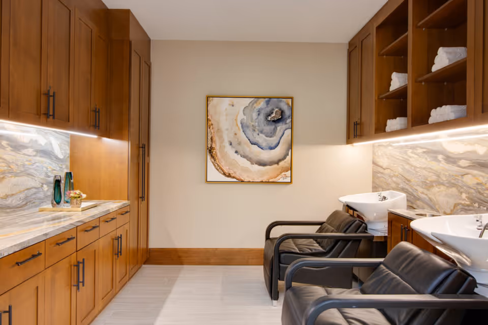 Interior view of a salon area in a senior living facility with two black salon chairs facing white wash basins. The room features wooden cabinets with black handles, marble countertops and backsplash, neatly stacked white towels on open shelves, and a framed abstract artwork on the wall.