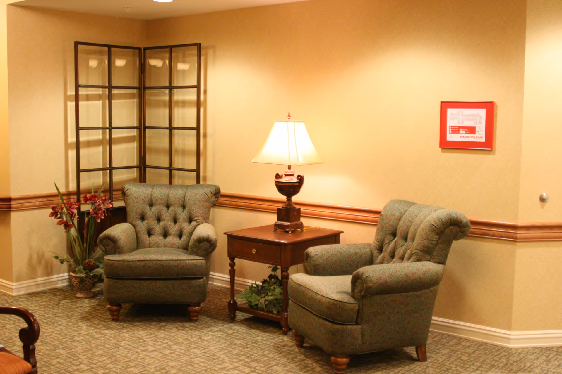 A cozy sitting area with two green upholstered armchairs facing each other, separated by a wooden side table with a lamp on it. There is a decorative folding screen in the corner and a plant with red flowers next to one of the chairs. The walls are beige with a wooden chair rail, and a framed evacuation plan is mounted on the wall.
