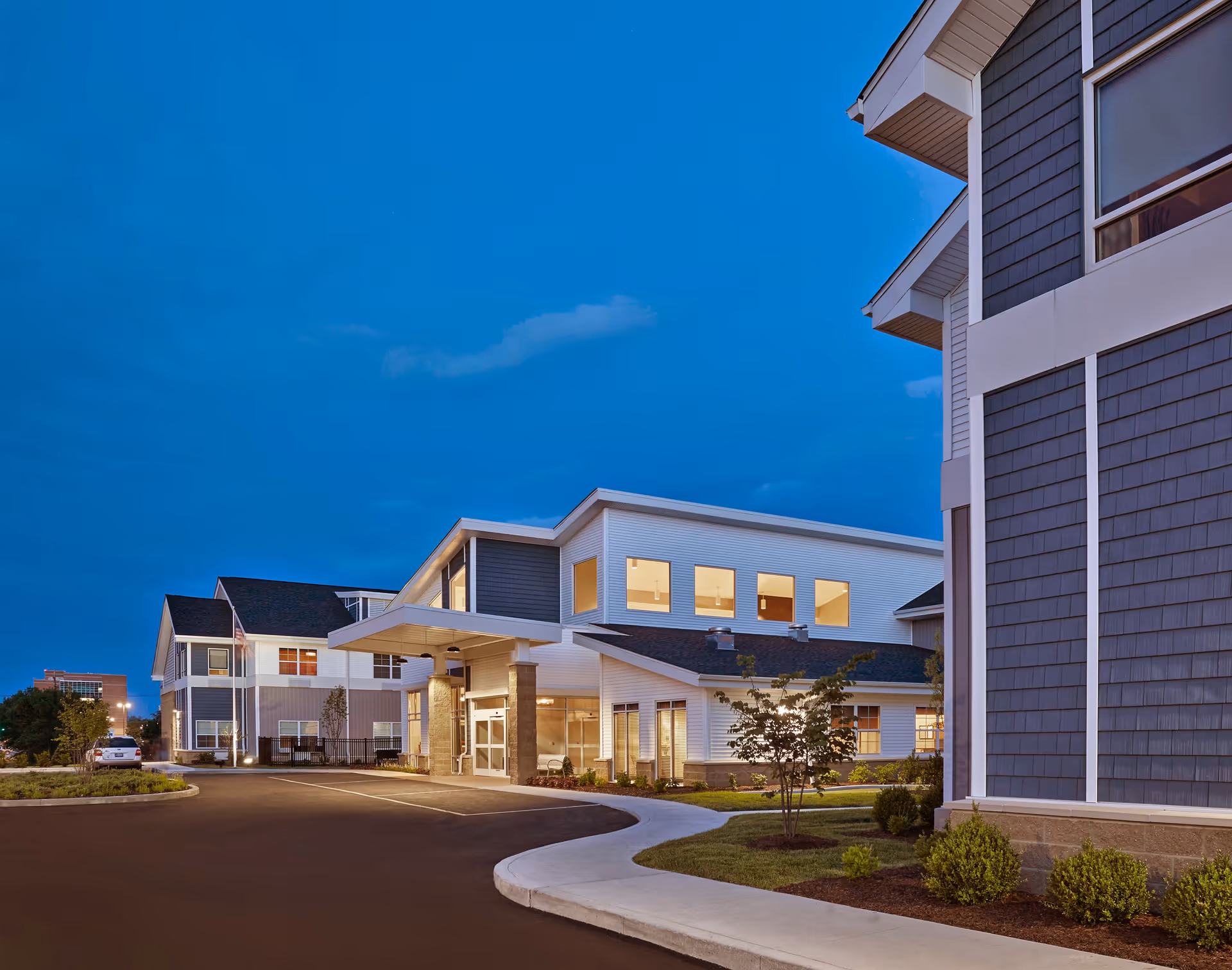 Exterior front view of a modern senior living facility at dusk with an illuminated entrance canopy, driveway and landscaped grounds.