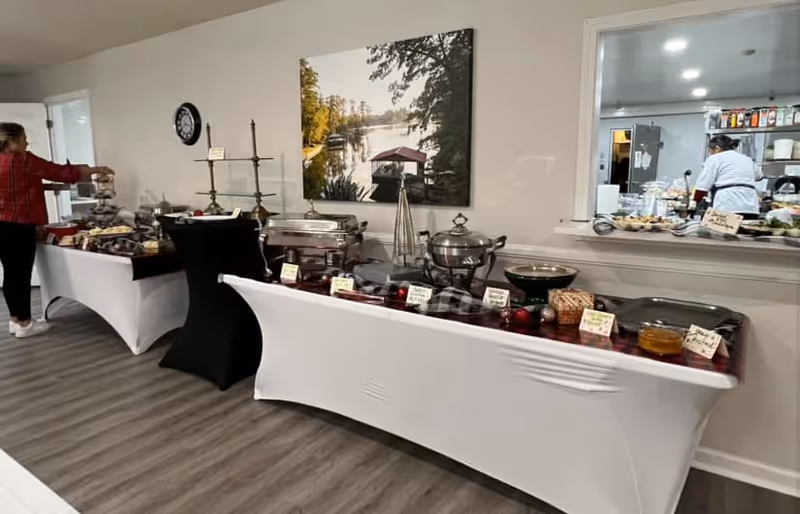 A buffet setup in a dining area with various covered dishes and food items on tables draped with white and black cloths. A woman is seen serving herself from the buffet on the left side. In the background, there is a large photograph of a lake scene on the wall and a kitchen window where a chef is visible preparing food.