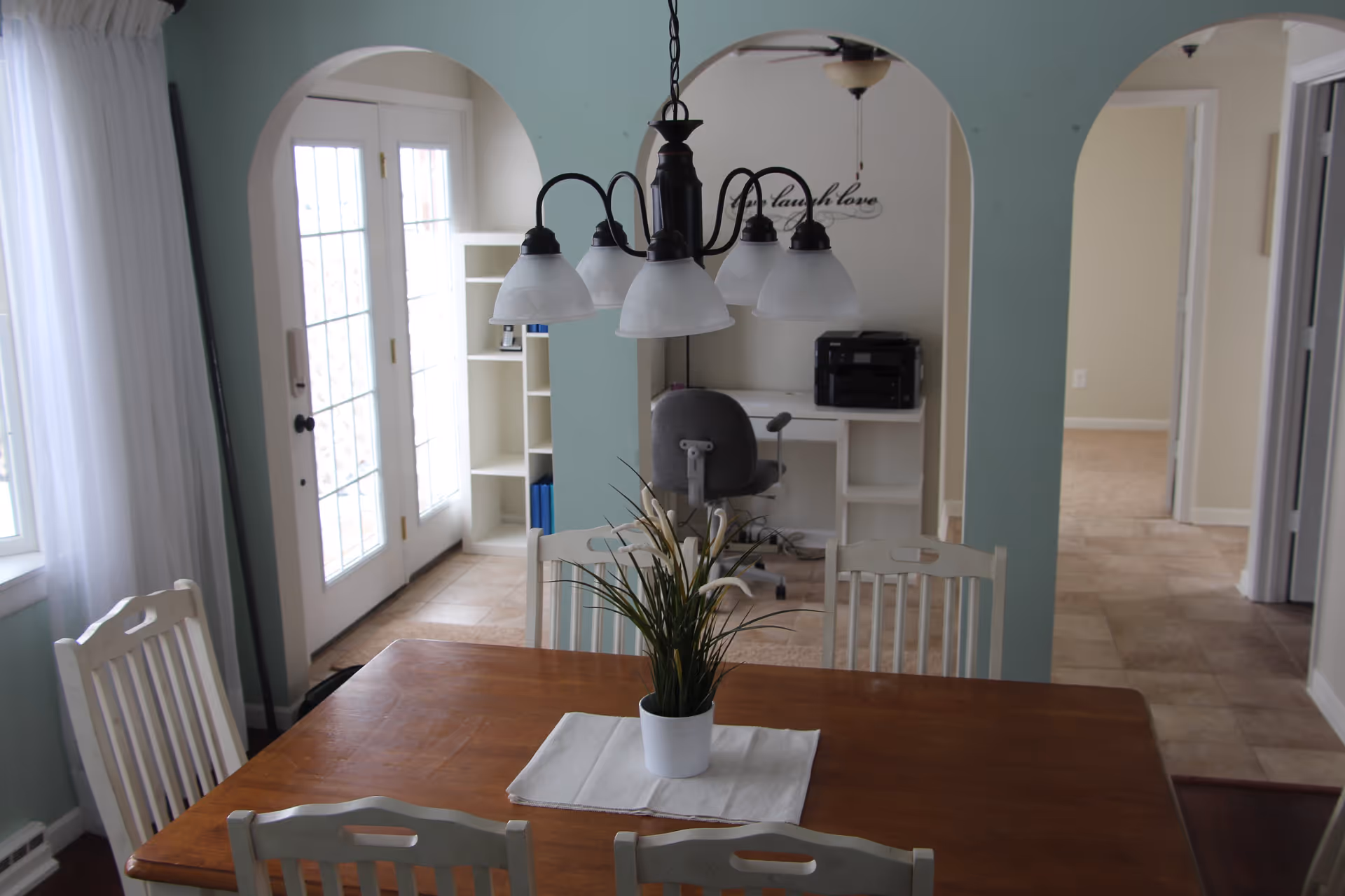 A dining area with a wooden table and white chairs. A white potted plant is centered on the table on a white placemat. Above the table hangs a black chandelier with five frosted glass shades. In the background, there is a light blue wall with two arched openings leading to a small office space with a gray office chair, white shelves, and a printer. To the left, there are glass double doors with white curtains, allowing natural light into the room. The floor is tiled in the background and wooden in the dining area.