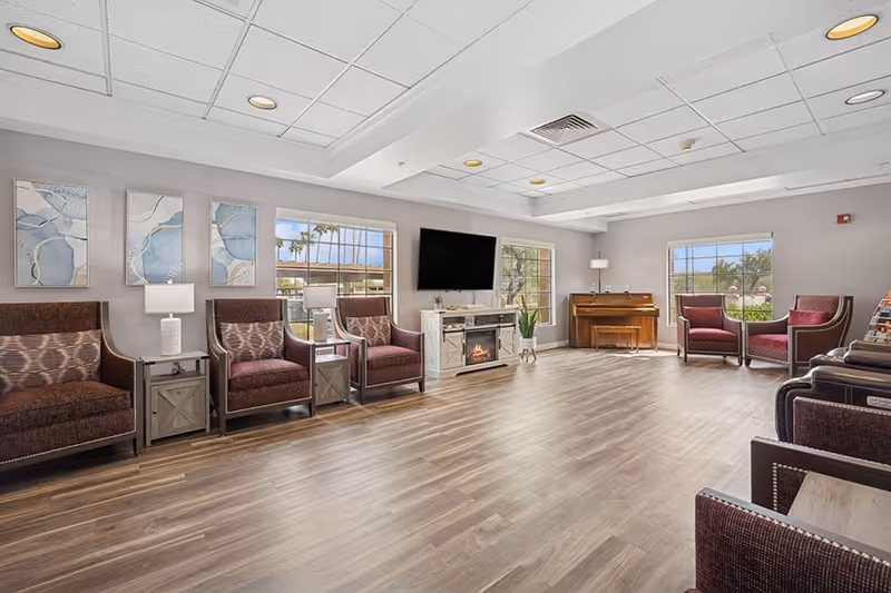 A spacious and well-lit common area with multiple armchairs arranged along the walls, a flat-screen TV mounted above an electric fireplace, a piano in the corner, and large windows letting in natural light. The room has wood flooring, neutral-colored walls, and modern ceiling lights.
