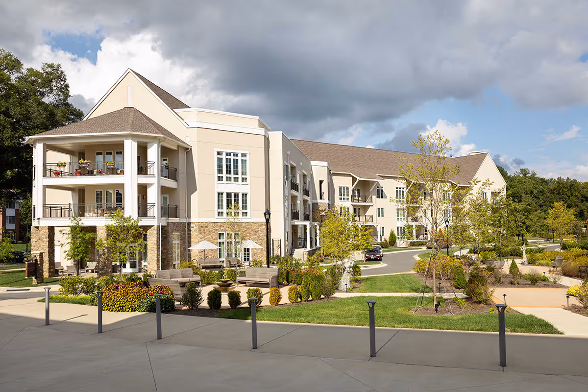 Exterior view of a multi-story senior living facility named Aldersgate, featuring balconies with outdoor furniture, landscaped gardens with trees and shrubs, paved walkways, and outdoor seating areas under umbrellas. The sky is partly cloudy with patches of blue.