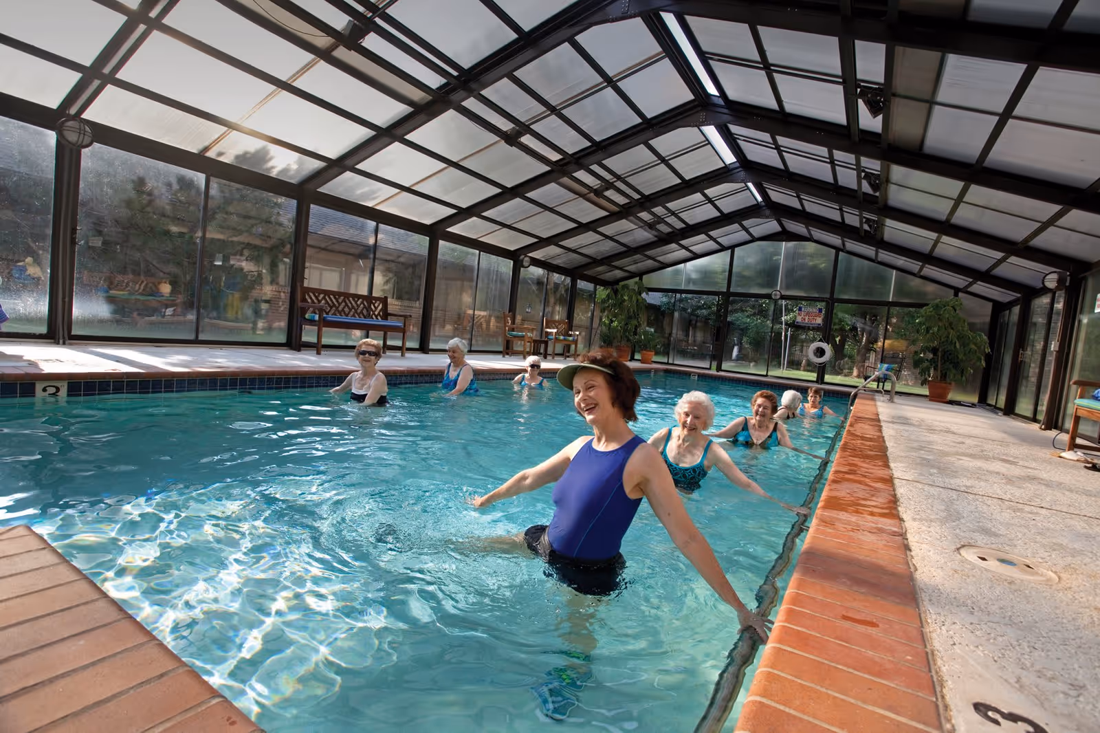 A group of elderly women participating in a water exercise class in an indoor swimming pool with a glass roof and walls. The women are smiling and appear to be enjoying the activity.