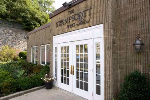 Exterior view of a building entrance with white double doors and glass panels. Above the doors is a sign that reads 'THE SWAMPSCOTT VING'. The building has a textured brown facade with a stone wall and greenery on the left side.