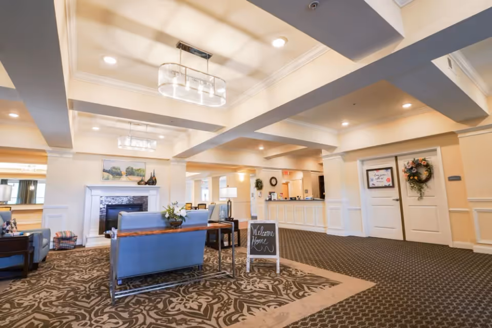 A spacious and well-lit senior living facility lobby with patterned carpet, blue upholstered seating, a fireplace with decorative items above it, a small table with a flower arrangement, and a chalkboard sign that reads 'Welcome Home'. The reception desk is visible in the background, along with double doors decorated with a wreath and a framed sign.