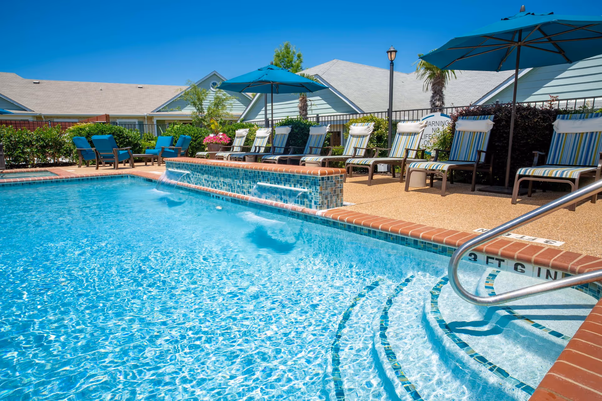 Outdoor swimming pool with clear blue water, surrounded by lounge chairs with striped cushions and umbrellas. There are some green bushes and residential-style buildings in the background under a clear blue sky.