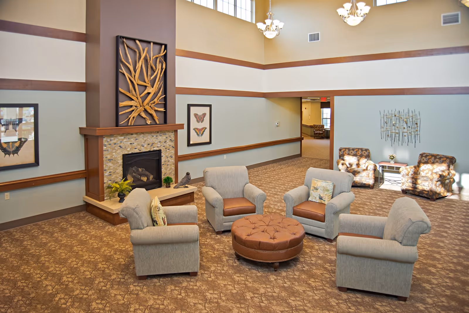 A spacious living room area with four beige armchairs arranged around a round brown ottoman. The room features a fireplace with a decorative wooden art piece above it, butterfly artwork on the walls, and two patterned armchairs near a small table with a flower vase. The room has high ceilings with large windows and hanging light fixtures.