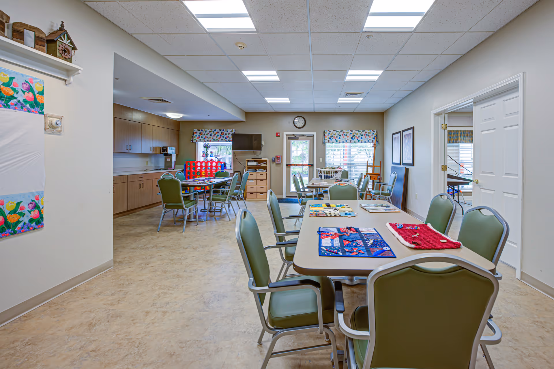 Bright common activity/dining room with tables and green chairs set up for crafts, a kitchenette along the left wall, and windows at the far end.