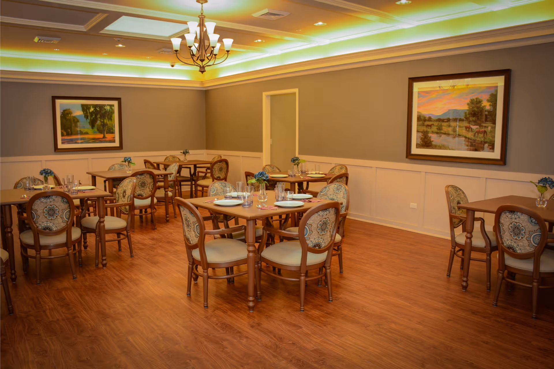 A dining room with several wooden tables and upholstered chairs arranged neatly. Each table is set with plates, glasses, and small flower vases. The room has wooden flooring, beige walls with white wainscoting, two framed landscape paintings, and a chandelier hanging from the ceiling.