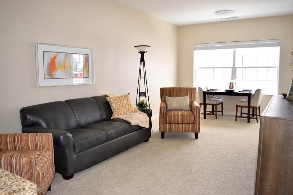 Bright living room with a black leather sofa, two striped armchairs, a tall floor lamp, and a small dining table by a large window.