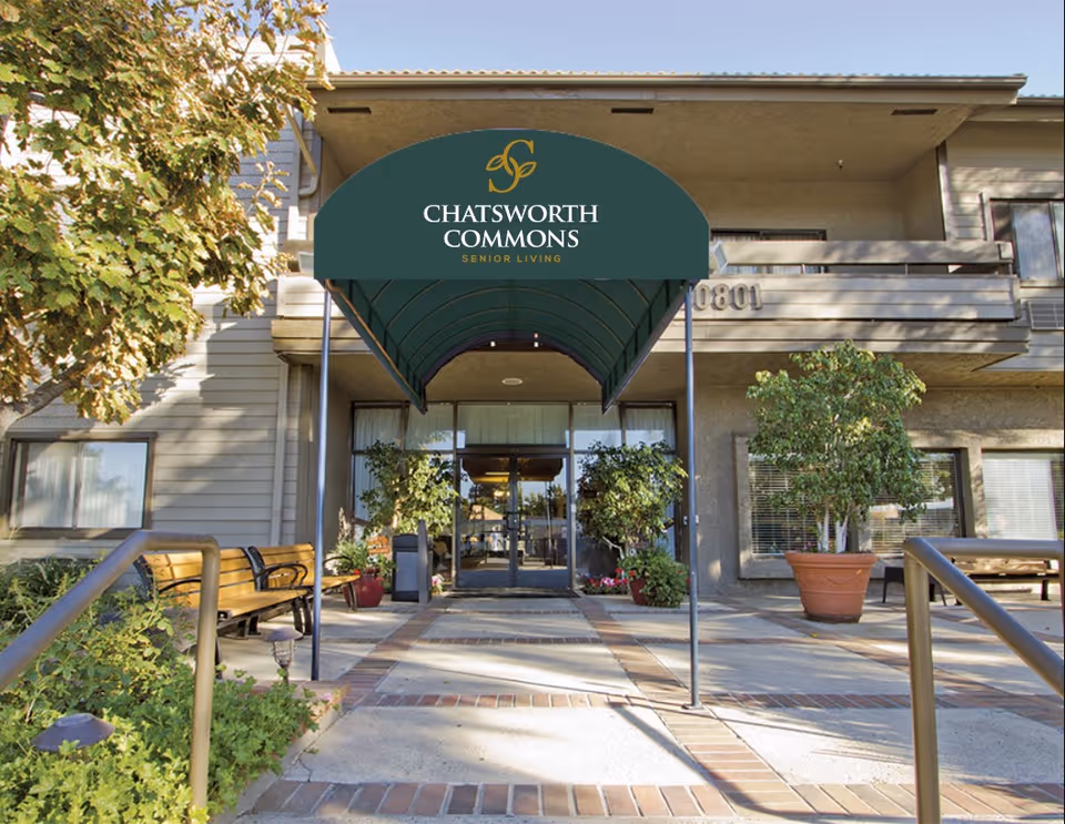 Front entrance of a senior living building with a green awning reading 'Chatsworth Commons', potted plants, benches and a paved walkway.
