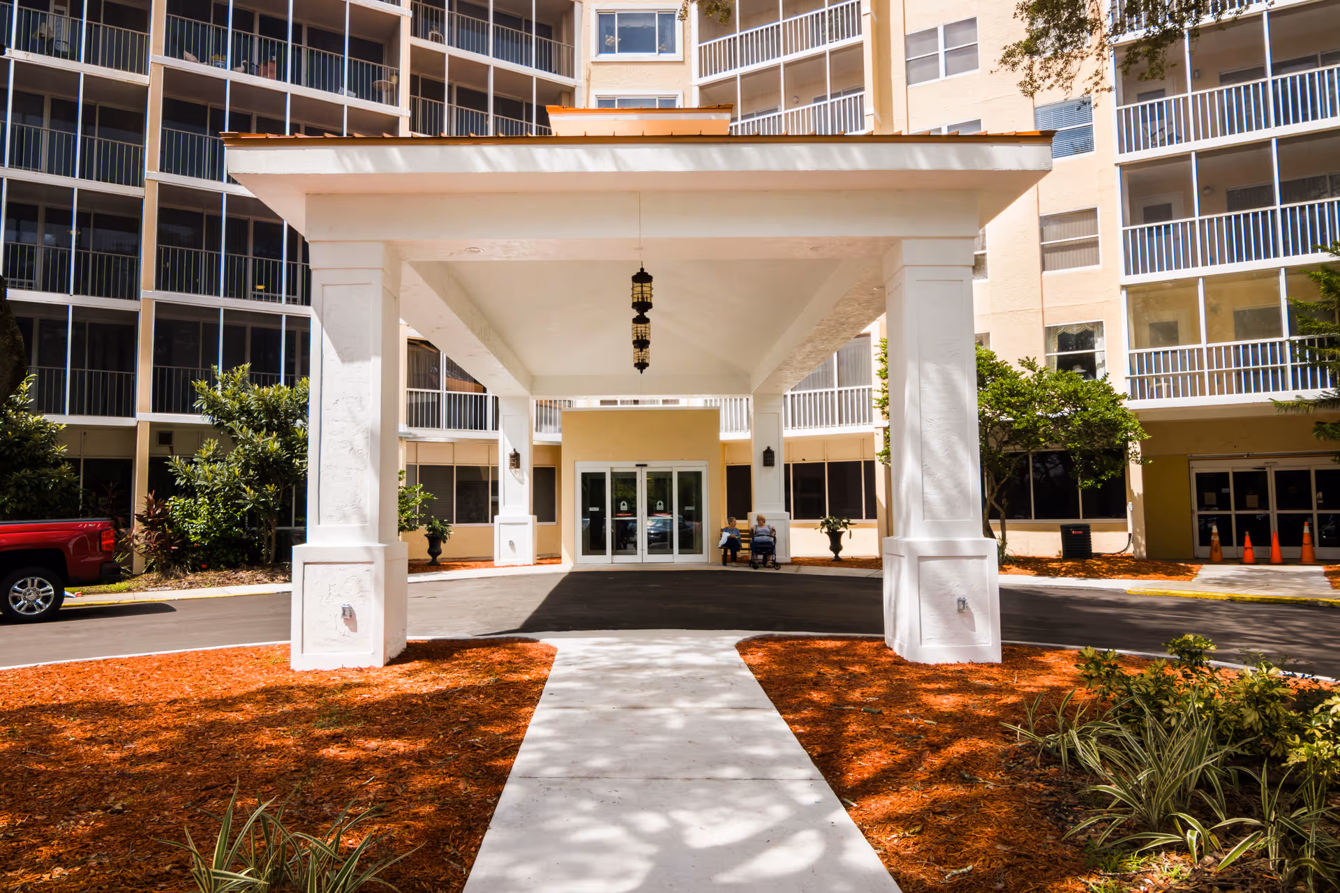 Entrance to a multi-story retirement community building with a covered drop-off area supported by white columns. Two people are sitting on a bench near the glass doors of the building. There are landscaped areas with mulch and plants on either side of the walkway leading to the entrance.
