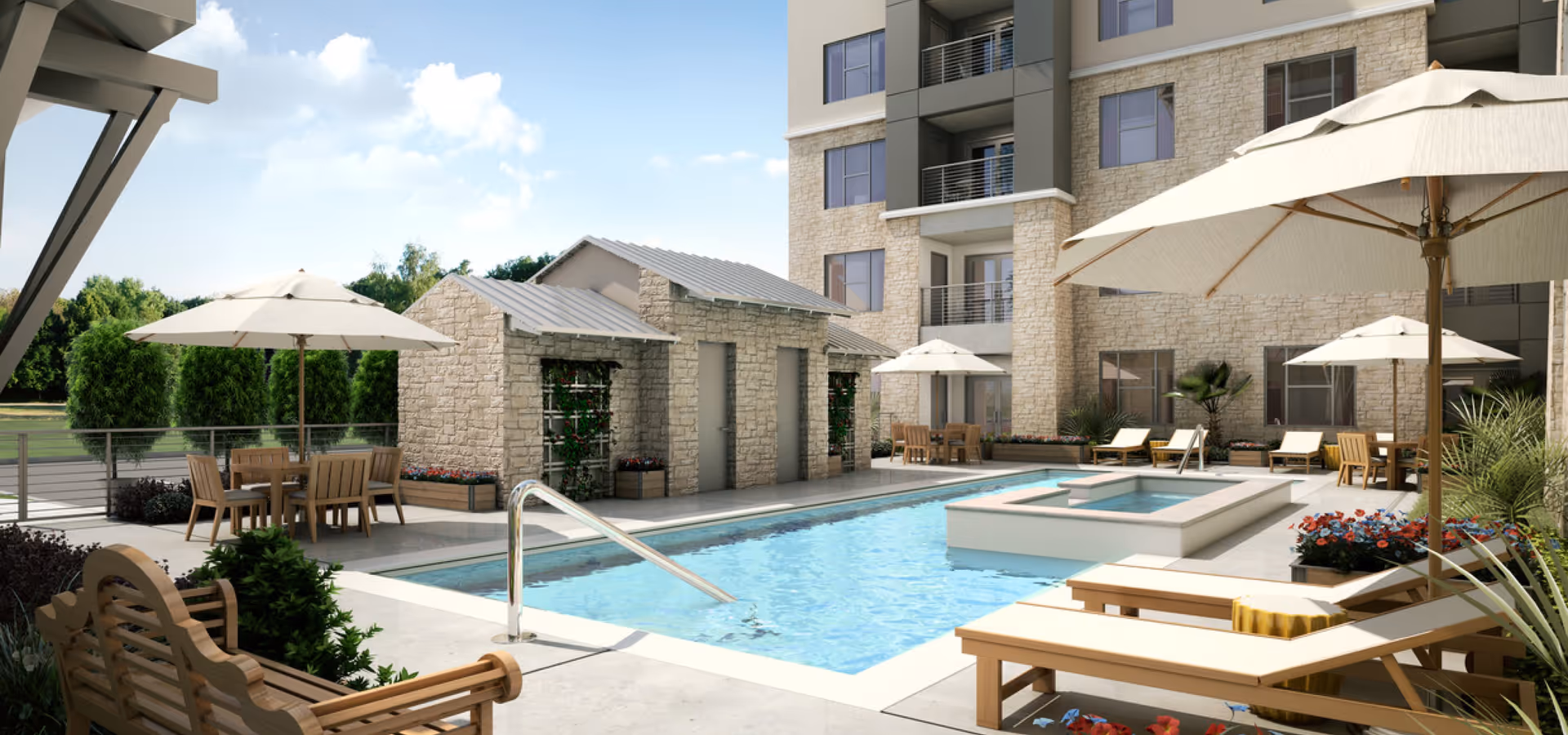 Outdoor pool area at Verena at Leander featuring a rectangular swimming pool with a built-in hot tub, surrounded by lounge chairs with cushions and umbrellas. There are tables with chairs and umbrellas for shade, stone buildings in the background, and a multi-story residential building behind the pool area. Greenery and trees are visible beyond the pool area.
