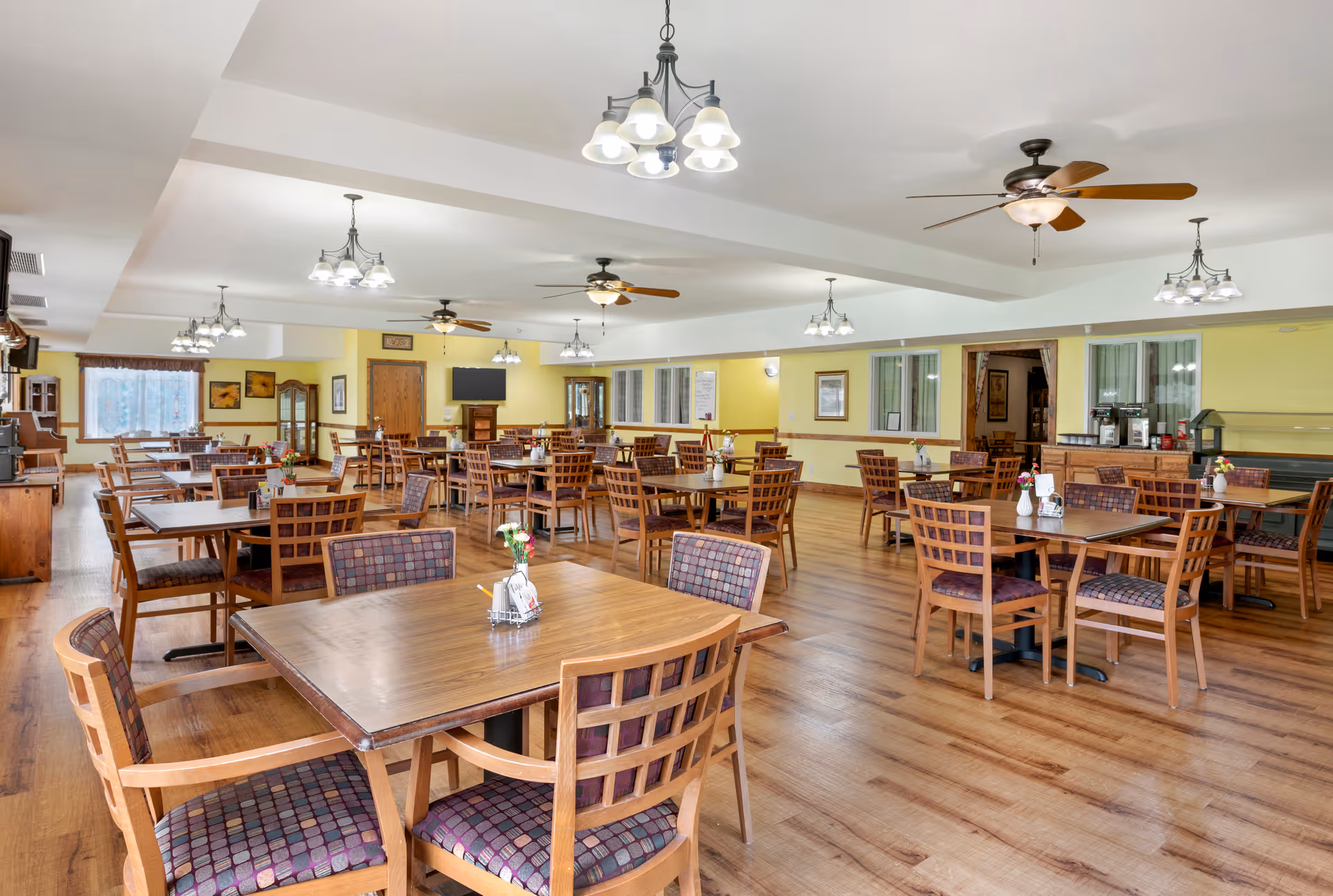 Spacious dining room with multiple wooden tables and chairs, ceiling fans and chandeliers, and a serving area in the back.