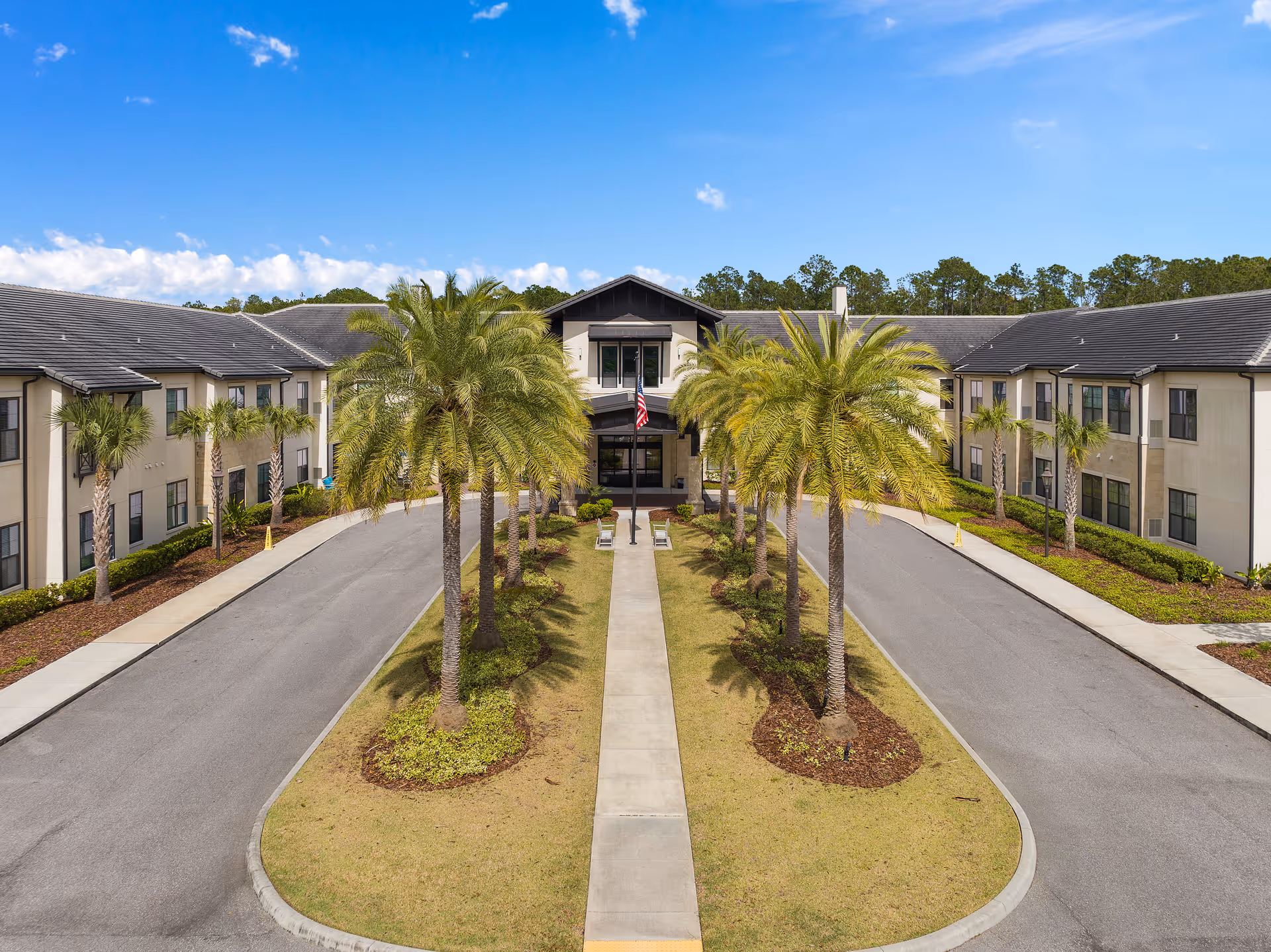 Front exterior view of Starling at Nocatee Assisted Living & Memory Care facility with a driveway lined by palm trees leading to the main entrance under a clear blue sky.