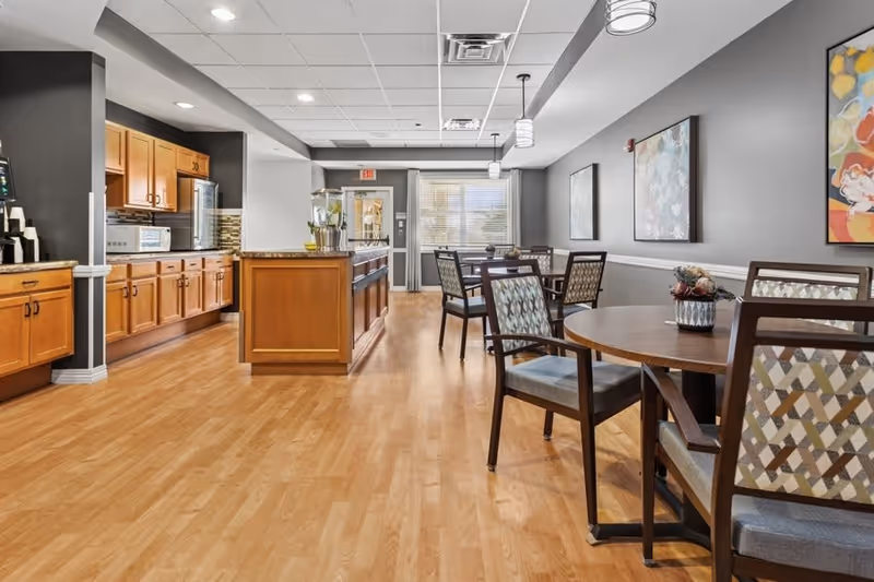 A bright dining area with wooden floors and multiple round tables surrounded by chairs with patterned upholstery. To the left, there is a kitchen area with wooden cabinets, a microwave, and a coffee station. The walls are painted gray and decorated with colorful abstract paintings. Large windows at the far end let in natural light.
