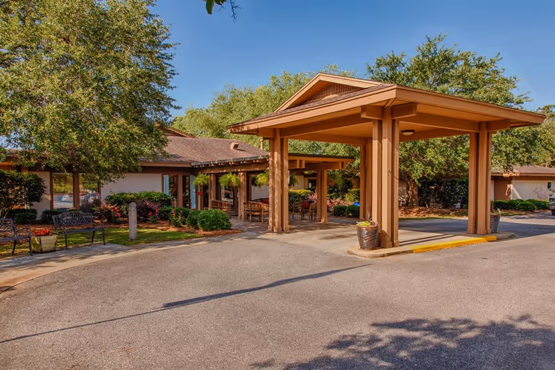Entrance area of The Brennity at Fairhope Senior Living with a covered drop-off structure, surrounded by trees, benches, and landscaped bushes under a clear blue sky.