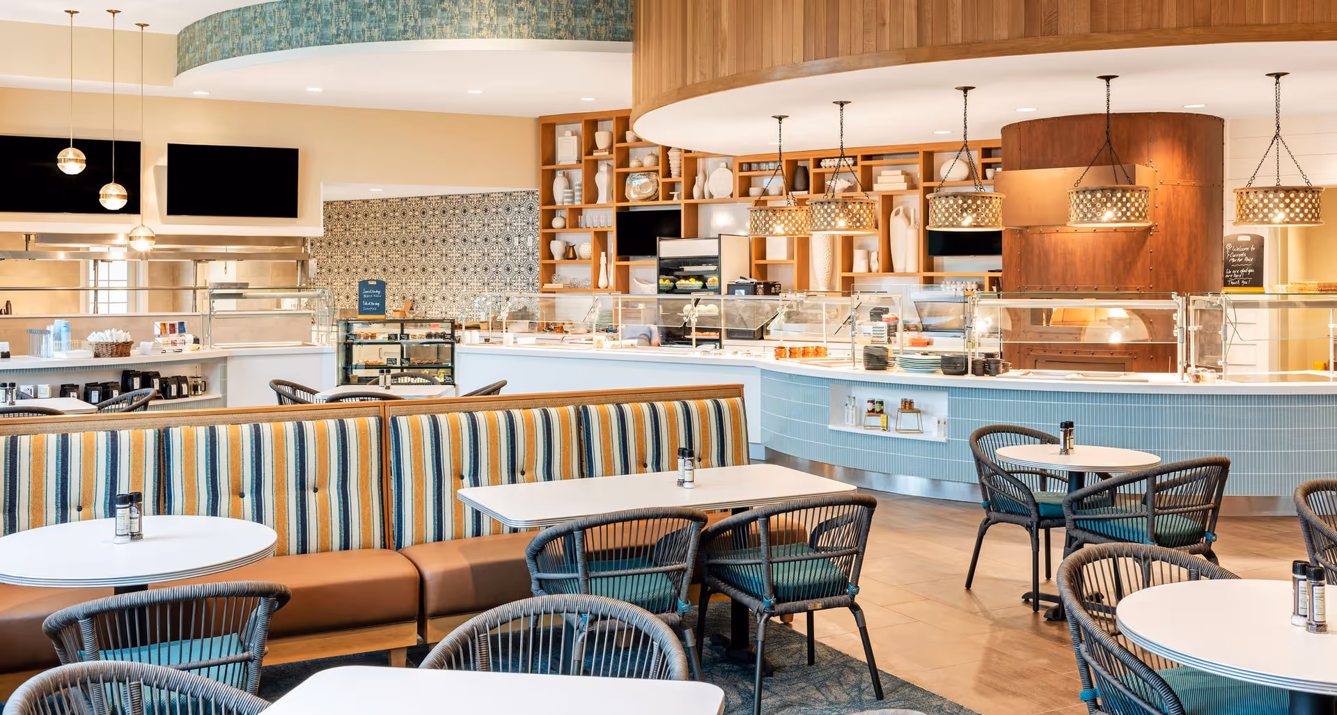 Bright modern dining area with round tables, wicker chairs, striped banquette seating, and a curved service counter with pendant lights.