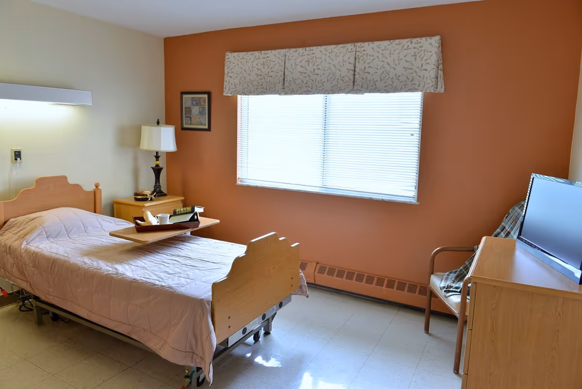 A simple senior living facility bedroom with a single bed covered in a light pink quilt. A wooden tray with a cup and a book is placed on the bed. Next to the bed is a wooden nightstand with a lamp and a small decorative item. The room has a large window with blinds and a patterned valance. Across from the bed is a wooden dresser with a flat-screen TV on top and a chair with a plaid blanket draped over it. The walls are painted in a combination of beige and orange tones.