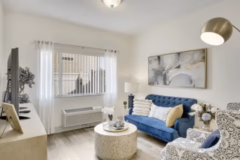 A bright living room with a blue tufted sofa, patterned armchair, round woven coffee table, and a TV on a light wood stand. The room has a large window with vertical blinds and sheer white curtains, a floor lamp, a table lamp, and abstract wall art above the sofa.