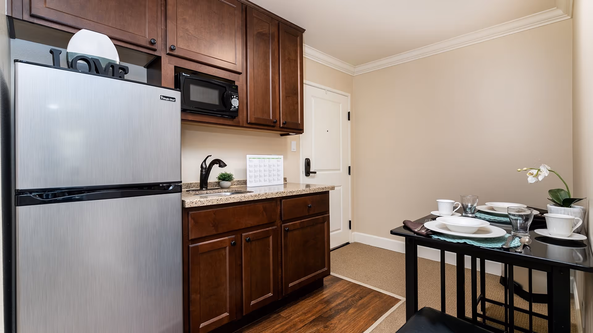Small kitchen area with dark wooden cabinets, a stainless steel refrigerator, a microwave, and a granite countertop. To the right, there is a small dining table set for two with white dishes, glasses, and a small potted orchid. The room has beige walls and a white door in the background.